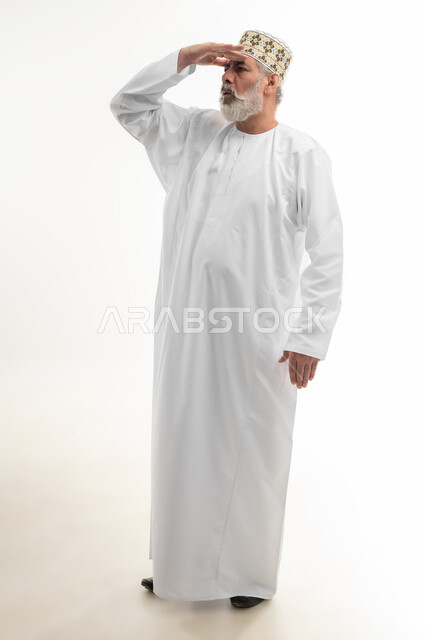 محاولة رؤية الأشياء البعيدة، إيماءات التركيز للحصول مشاهدة واضحة، بورتريه لرجل كبير بالسن عربي خليجي عماني يرتدي الدشداشة التقليدية والكمه العمانية يضع يده على جبينه وينظر إلى اتجاه معين، الجسم بالطول الكامل، خلفية بيضاء