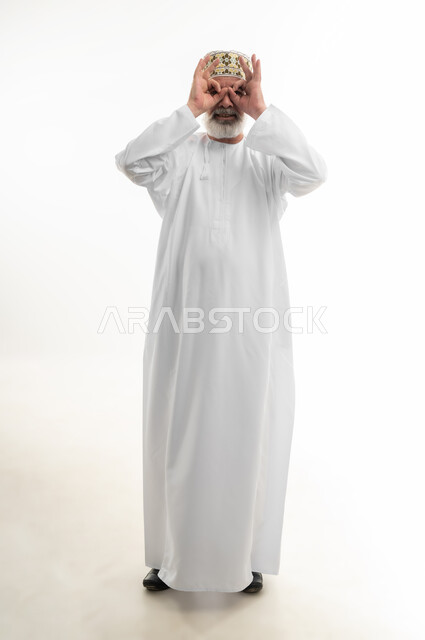 التخطيط والرؤية المستقبلية، وضع اليدين على العينين وتمثيل حركة المنظار، بورتريه لرجل كبير بالسن عربي خليجي عماني يرتدي الدشداشة التقليدية والكمه العمانية وينظر إلى الكاميرا بإيماءات البشاشة، التأمل بتحقيق الأحلام والطموحات، الجسم بالطول الكامل، خلفية بيضاء