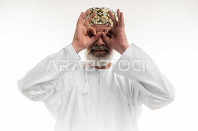 التأمل بتحقيق الأحلام والطموحات، وضع اليدين على العينين وتمثيل حركة المنظار، بورتريه مقرب لرجل كبير بالسن عربي خليجي عماني يرتدي الدشداشة التقليدية والكمه العمانية وينظر إلى الكاميرا بإيماءات السرور والبشاشة، رؤية الخطط المستقبلية، خلفية بيضاء