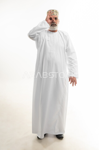 رؤية الخطط المستقبلية، التأمل بتحقيق الأحلام والطموحات، وضع اليد على إحدى العينين وتمثيل حركة المنظار الأحادي، بورتريه لرجل كبير بالسن عربي خليجي عماني يرتدي الدشداشة التقليدية والكمه العمانية وينظر للكاميرا بإيماءات السرور، الجسم بالطول الكامل، خلفية بيضاء