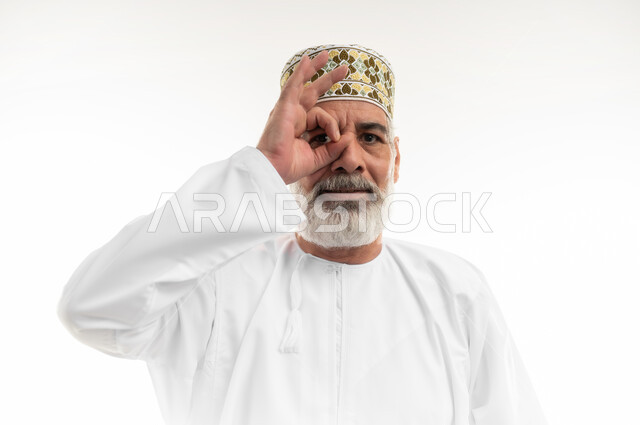تمثيل حركة المنظار الأحادي بوضع اليد على إحدى العينين، رؤية الخطط المستقبلية، التأمل بتحقيق الأحلام والطموحات، بورتريه مقرب لرجل كبير بالسن عربي خليجي عماني يرتدي الدشداشة التقليدية والكمه العمانية وينظر للكاميرا بإيماءات السرور، خلفية بيضاء