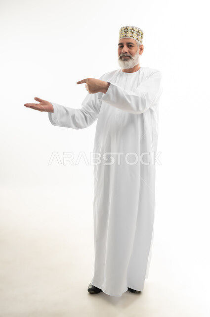 Looking at camera with gestures of pleasure and pointing finger at something, portrait of an old Arab Gulf Omani man wearing a dishdasha and traditional Omani kummah standing straight and holding an imaginary item in his hand, interest in appearance, full body, white background
