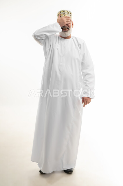 إيماءات تذكر شيء ما بشكل مفاجئ، حركات اليدين والوجه تعبر عن الخجل، بورتريه لرجل شايب عربي خليجي عماني يرتدي الدشداشة والكمه العمانية التقليدية يقف بشكل مستقيم ويغطي عينيه بيده، الاهتمام بالمظهر الخارجي، الجسم بالطول الكامل، خلفية بيضاء