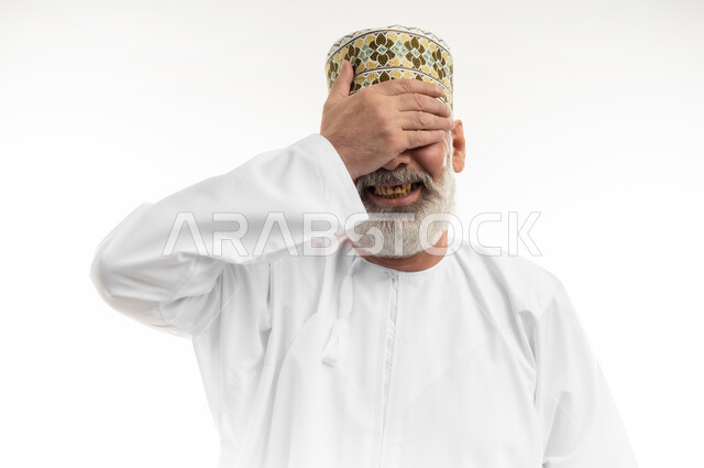 Hand and face movements expressing shyness, gestures that suddenly remember something, close-up portrait of an old Arab Gulf Omani man wearing a dishdasha and traditional Omani kummah standing straight and covering his eyes with his hand, concern for outward appearance, white background