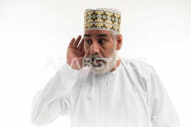 إيماءات الدهشة والاستغراب بسماع شيء ما صادم، محاولة استراق السمع، ضعف القدرة على الاستماع، بورتريه مقرب لرجل شايب عربي خليجي عماني يرتدي الدشداشة والكمه العمانية التقليدية يقف بشكل مستقيم ويضع يده خلف أذنه بتعابير المفاجأة، خلفية بيضاء