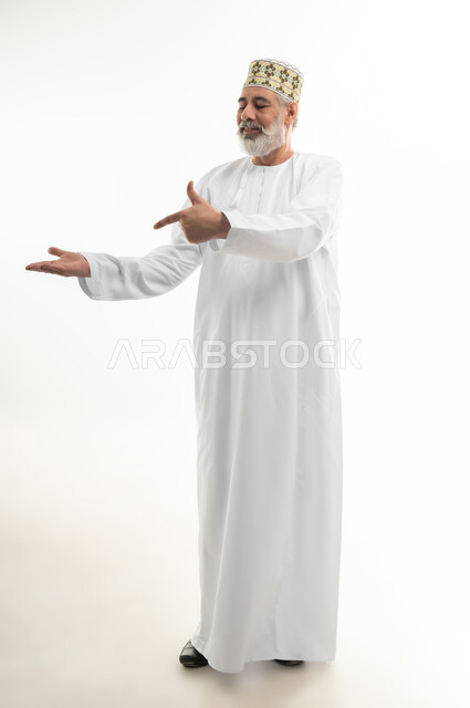الإشارة بالاصبع إلى شيء ما بإيماءات السرور، بورتريه لرجل شايب عربي خليجي عماني يرتدي الدشداشة والكمه العمانية التقليدية يقف بشكل مستقيم ويحمل بيده عنصر وهمي، الاهتمام بالمظهر الخارجي، الجسم بالطول الكامل، خلفية بيضاء
