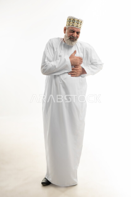 آلام الذبحة الصدرية، التعرض لنوبات قلبية مفاجئة، الإحساس بوخزة في الصدر والقلب، بورتريه لرجل شايب عربي خليجي عماني يرتدي الدشداشة والكمه العمانية التقليدية يضع يده على قلبه بإيماءات التوجع والألم، الجسم بالطول الكامل، خلفية بيضاء