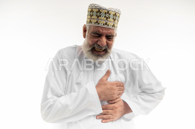 التعرض لنوبات قلبية مفاجئة، آلام الذبحة الصدرية، الإحساس بوخزة في الصدر والقلب، بورتريه مقرب لرجل شايب عربي خليجي عماني يرتدي الدشداشة والكمه العمانية التقليدية يضع يده على قلبه بإيماءات التوجع والألم، خلفية بيضاء