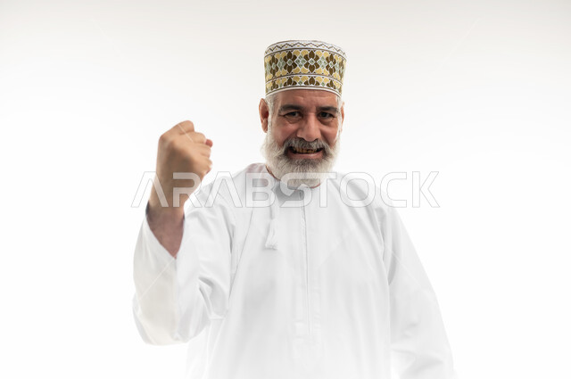 التشجيع بتعابير الحماس، الفرح بالانتصار والفوز، رفع قبضة اليد نحو الأعلى بإيماءات القوة والتحدي، بورتريه مقرب لرجل شايب عربي خليجي عماني يرتدي الدشداشة والكمه العمانية التقليدية يرفع يده في الهواء بسعادة وسرور، خلفية بيضاء