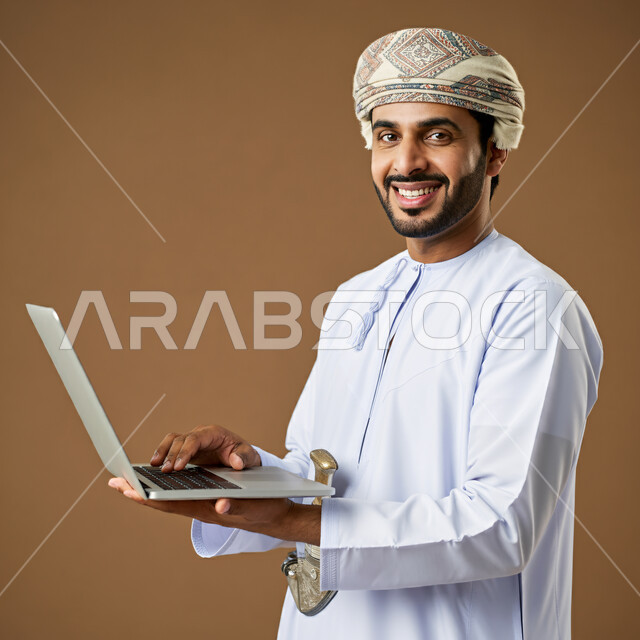 مفهوم تسهيل الاعمال والقدرة على إنجاز المهام عن بعد، استخدام التقنيات الحديثة والأجهزة المتطورة ، بورتريه لشاب عربي خليجي عماني يرتدي الدشداشة والعمامة يمسك بيده حاسوب محمول تظهر عليه علامات الفرح والسرور، خلفية بيج