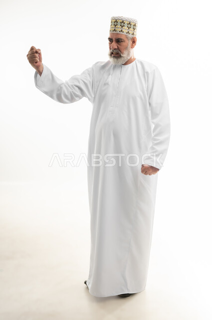رفع قبضة اليد نحو الأعلى بإيماءات القوة والتحدي، التشجيع بتعابير الحماس، الفرح بالانتصار والفوز، بورتريه لرجل شايب عربي خليجي عماني يرتدي الدشداشة والكمه العمانية التقليدية يرفع يده في الهواء بسعادة وسرور، الجسم بالطول الكامل، خلفية بيضاء