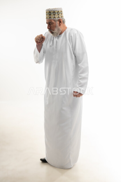Symptoms of Corona virus and the need for medical advice, cough and chest infections, portrait of an old Arab Gulf Omani man wearing a dishdasha and the traditional Omani kummah, putting his hand in front of his mouth and suffering from a severe cough, full body, white background