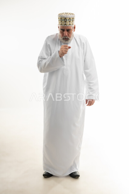 السعال والالتهابات الصدرية، ظهور أعراض مرض كورونا والحاجة للاستشارة الطبية، بورتريه لرجل شايب عربي خليجي عماني يرتدي الدشداشة والكمه العمانية التقليدية يضع يده أمام فمه ويعاني من الكحة القوية، الجسم بالطول الكامل، خلفية بيضاء
