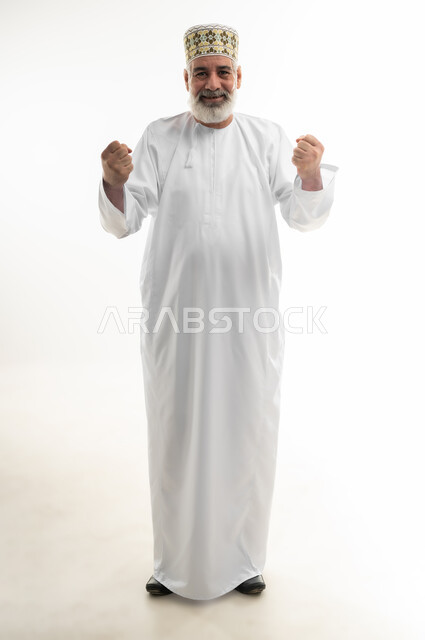 تحقيق الإنجازات والوصول إلى الأهداف والطموحات، قبض ورفع اليدين نحو الأعلى والتشجيع بحماس، الفرح بالنصر والفوز، بورتريه لرجل شايب عربي خليجي عماني يرتدي الدشداشة والكمه العمانية التقليدية ينظر للكاميرا بإيماءات السعادة، الجسم بالطول الكامل، خلفية بيضاء