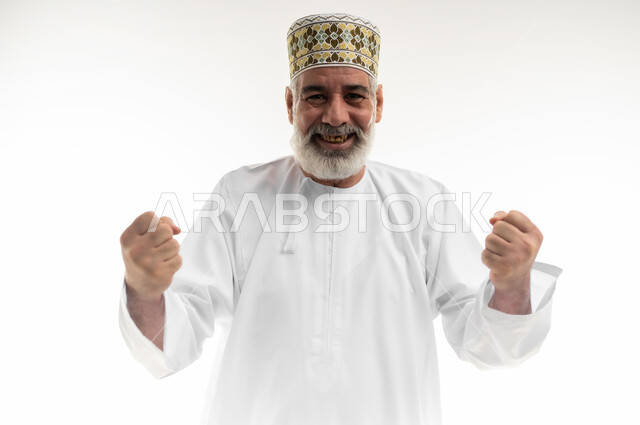 Joy of victory and triumph, achieving accomplishments and reaching goals and ambitions, clenching and raising hands upwards and cheering enthusiastically, close-up portrait of an old Arab Gulf Omani man wearing a dishdasha and traditional Omani kummah looking at the camera with gestures of happiness, white background