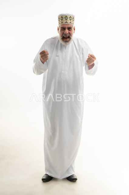 قبض ورفع اليدين نحو الأعلى والتشجيع بحماس، تحقيق الإنجازات والوصول إلى الأهداف والطموحات، الفرح بالنصر والفوز، بورتريه لرجل شايب عربي خليجي عماني يرتدي الدشداشة والكمه العمانية التقليدية ينظر للكاميرا بإيماءات السعادة، الجسم بالطول الكامل، خلفية بيضاء