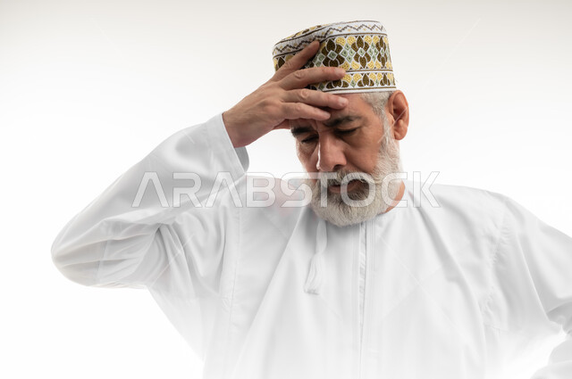 التعرض لوعكة صحية، الشعور بالتعب والإرهاق، إغماض العينين والإحساس بالدوار، بورتريه مقرب لرجل شايب عربي خليجي عماني يرتدي الدشداشة والكمه العمانية التقليدية يضع يده على رأسه بتعابير الاضطراب والخمول، خلفية بيضاء
