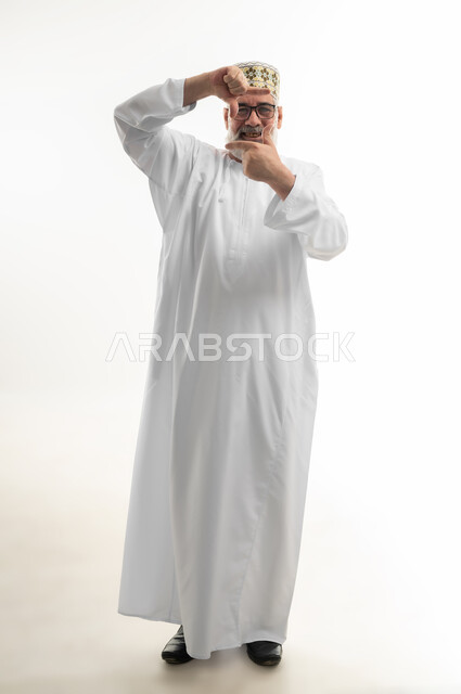 Using hands to make a rectangular frame, trying to size something, portrait of an old Arab Gulf Omani man wearing a dishdasha, a sleeve and glasses looking at the camera with gestures of happiness and pleasure, full body, white background
