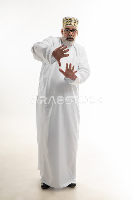 Panic and request to stop and leave a safe distance, panic and fear of something, extending hands forward in an attempt to defend oneself, portrait of an old Arab Gulf Omani man wearing a dishdasha, a sleeve and glasses and looking at the camera with gestures of terror and awe, full body, white background
