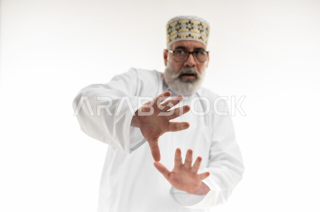 Panic and fear of something, hands extended forward in an attempt to defend oneself, panic and request to stop and leave a safe distance, close-up portrait of an old Arab Gulf Omani man wearing a dishdasha, a sleeve and glasses and looking at the camera with gestures of terror and awe, white background