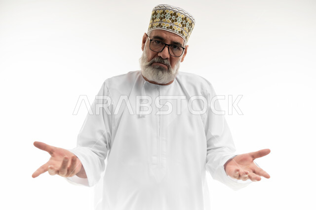 التردد وعدم التأكد بشأن أمر معين، فتح اليدين في حيرة وعدم القدرة على اتخاذ القرار، بورتريه مقرب لرجل شايب عربي خليجي عماني يرتدي الدشداشة والكمه والنظارة الطبية يرفع كفيه نحو الأعلى وينظر إلى الكاميرا بإيماءات الاستغراب، خلفية بيضاء