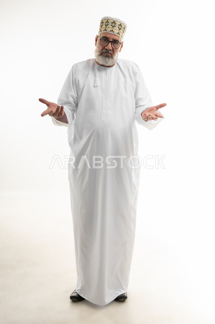 فتح اليدين في حيرة وعدم القدرة على اتخاذ القرار، التردد وعدم التأكد بشأن أمر معين، بورتريه لرجل شايب عربي خليجي عماني يرتدي الدشداشة والكمه والنظارة الطبية يرفع كفيه نحو الأعلى وينظر إلى الكاميرا بإيماءات الاستغراب، الجسم بالطول الكامل، خلفية بيضاء