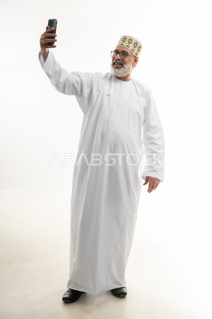 Taking selfies with a mobile phone, using modern devices and technology to immortalize memories, portrait of an old Arab Gulf Omani man wearing a dishdasha, a sleeve, and glasses, holding a mobile phone and taking selfies with gestures of pleasure, full body, white background