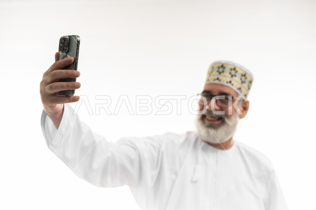 Using modern devices and technology to immortalize memories, taking beautiful pictures using a mobile phone, a close-up portrait of an old Arab Gulf Omani man wearing a dishdasha, a sleeve, and medical glasses, holding a mobile phone and taking selfies with gestures of happiness and pleasure, white background