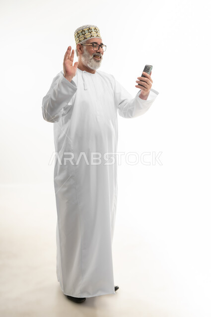 التواصل مع الأهل والأصدقاء وسماع أخبارهم، الاستفادة من الأجهزة والتكنولوجيا الحديثة في الاطمئنان على الأقارب، بورتريه لرجل شايب عربي خليجي عماني يرتدي الدشداشة والكمه والنظارة الطبية ويمسك الهاتف المحمول بإيماءات السعادة، الجسم بالطول الكامل، خلفية بيضاء