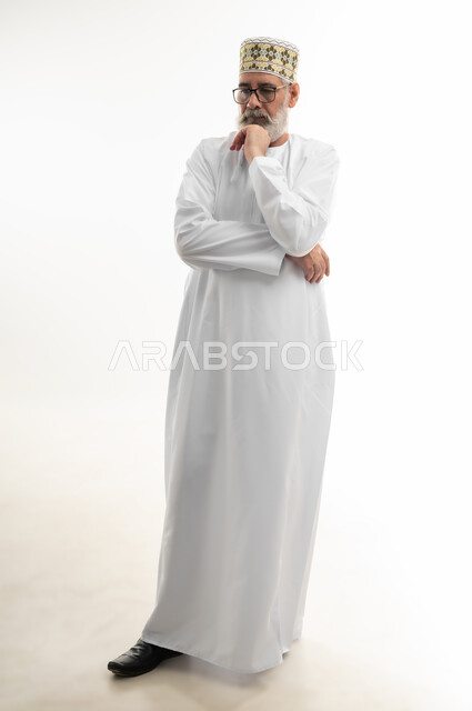 الحيرة في اتخاذ القرار وحل المشكلات، النظر إلى مكان ما بشرود الذهن، الإحساس بالملل والضجر، بورتريه لرجل شايب عربي خليجي عماني يرتدي الدشداشة والكمه العمانية ويلبس النظارة الطبية يضع يده أسفل ذقنه بإيماءات التفكير العميق، الجسم بالطول الكامل، خلفية بيضاء