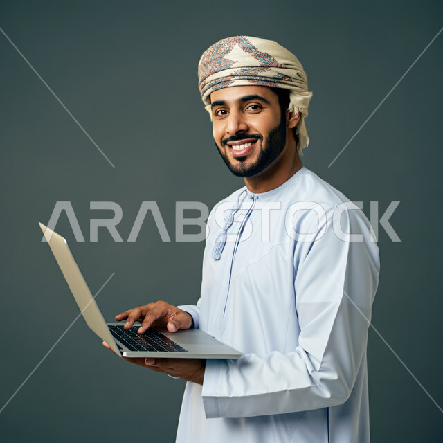  مفهوم تسهيل الاعمال والقدرة على إنجاز المهام عن بعد، استخدام التقنيات الحديثة والأجهزة المتطورة ، بورتريه لشاب عربي خليجي عماني يرتدي الدشداشة والعمامة يمسك بيده حاسوب محمول تظهر عليه علامات الفرح والسرور، خلفية رمادية