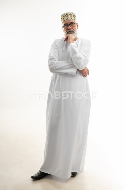 النظر إلى مكان ما بشرود الذهن، الإحساس بالملل والضجر، الحيرة في اتخاذ القرار وحل المشكلات، بورتريه لرجل شايب عربي خليجي عماني يرتدي الدشداشة والكمه العمانية ويلبس النظارة الطبية يضع يده أسفل ذقنه بإيماءات التفكير العميق، الجسم بالطول الكامل، خلفية بيضاء