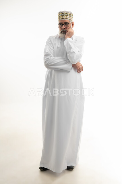 الإحساس بالملل والحاجة لتغيير الروتين اليومي، النظر إلى مكان ما بشرود، بورتريه لرجل شايب عربي خليجي عماني يرتدي الدشداشة والكمه العمانية ويلبس النظارة الطبية يضع يده على خده بإيماءات التفكير العميق، الجسم بالطول الكامل، خلفية بيضاء