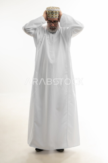 أعراض الصداع النصفي، نوبات الشقيقة الحادة، بورتريه لرجل شايب عربي خليجي عماني يرتدي الدشداشة والكمه العمانية يمسك رأسه بيديه بإيماءات التعب والألم الشديد، آلام الرأس والحاجة لأخذ المسكنات والراحة، الجسم بالطول الكامل، خلفية بيضاء
