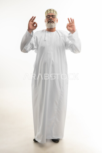 رفع اليدين نحو الأعلى بحركات تدل على القبول والامتياز، بورتريه لرجل شايب عربي خليجي عماني يرتدي الدشداشة والكمه العمانية ينظر إلى الكاميرا بإيماءات البشاشة والسرور، علامة الجودة التامة، تلقي الاستحسان بشكل كبير، الجسم بالطول الكامل، خلفية بيضاء
