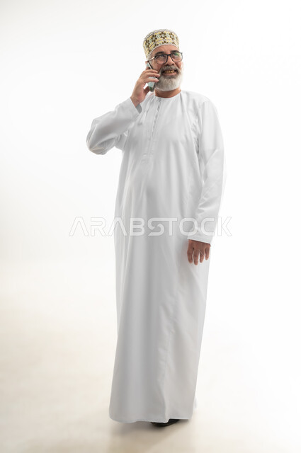 إيماءات السعادة بسماع أخبار جيدة، استخدام التقنيات والتكنولوجيا الحديثة والمتطورة، بورتريه لرجل شايب عربي خليجي عماني يرتدي الدشداشة والكمه العمانية ويلبس النظارة الطبية يمسك الهاتف المحمول بيده ويجري مكالمة هاتفية، الجسم بالطول الكامل، خلفية بيضاء