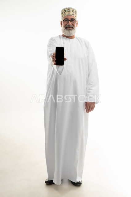 عرض شاشة جوال سوداء فارغة، استخدام التقنيات والتكنولوجيا الحديثة والمتطورة، بورتريه لرجل شايب عربي خليجي عماني يرتدي الدشداشة والكمه العمانية ويلبس النظارة الطبية يمسك الهاتف المحمول بيده وينظر إلى الكاميرا بإيماءات البشاشة، الجسم بالطول الكامل، خلفية بيضاء