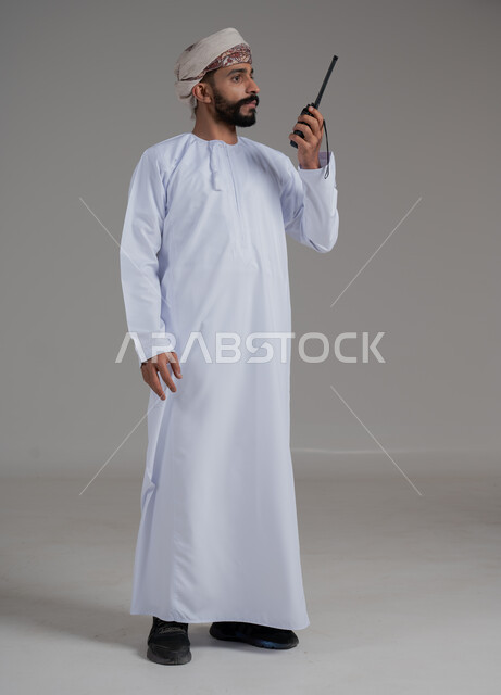 المتابعة والإشراف على خطط العمل بايماءات السعادة والبشاشة، بورتريه لرجل عربي خليجي عماني مبتسم يرتدي الدشداشة والعمامة يقوم باستخدم جهاز الارسال والاستقبال اللاسلكي، مفهوم ادارة المشاريع عن بعد، الإستجابة لحالات الطوارئ، صورة لكامل الجسم، خلفية رمادية