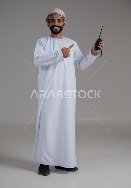 Pointing with index finger on the wireless device, portrait of an Arab Gulf Omani man wearing a dishdasha and turban using a wireless transmitter and receiver, giving work orders, using modern communication methods and technologies, responding to emergencies, full body length, gray background