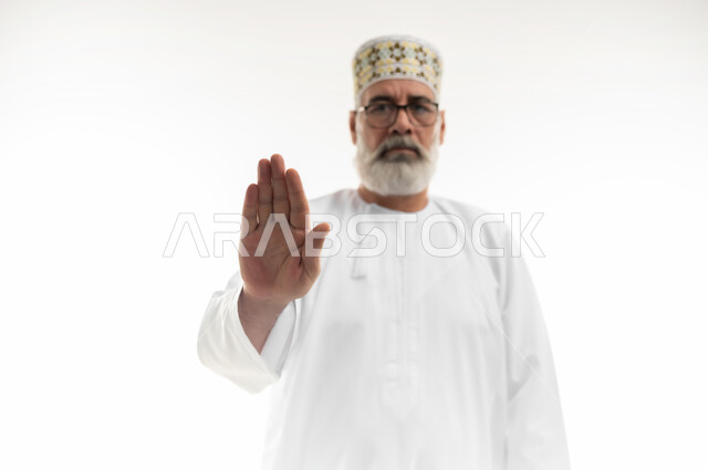 فتح الكف وتوجيهه نحو الأمام بتعابير الحزم والنهي، الأمر بالتوقف عن فعل شيء ما، طلب ترك مسافة بين الأشخاص، بورتريه مقرب لرجل كبير بالسن عربي خليجي عماني يرتدي الدشداشة والكمه التقليدية العمانية وينظر للكاميرا بإيماءات الغضب والجدية، خلفية بيضاء