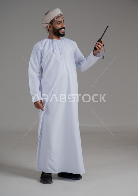 الإستجابة لحالات الطوارئ، المتابعة والإشراف على خطط العمل بايماءات السعادة والبشاشة، بورتريه لرجل عربي خليجي عماني مبتسم يرتدي الدشداشة والعمامة يقوم باستخدم جهاز الارسال والاستقبال اللاسلكي، مفهوم ادارة المشاريع عن بعد، صورة لكامل الجسم، خلفية رمادية