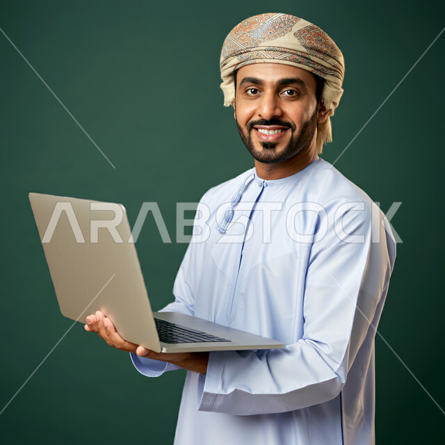  مفهوم تسهيل الاعمال والقدرة على إنجاز المهام عن بعد، استخدام التقنيات الحديثة والأجهزة المتطورة ، بورتريه لشاب عربي خليجي عماني يرتدي الدشداشة والعمامة يمسك بيده حاسوب محمول تظهر عليه علامات الفرح والسرور، خلفية خضراء