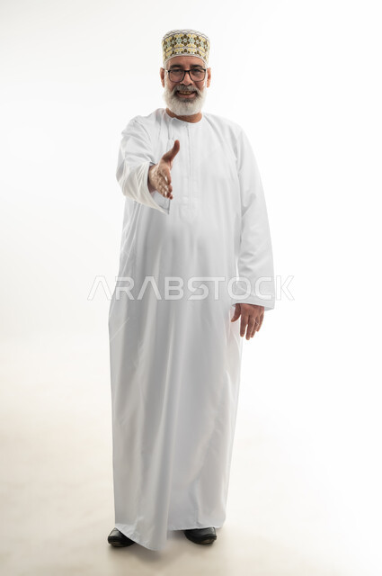 فتح كف اليد ومده نحو الأمام بنية السلام والمصافحة، حركات الوجه واليدين تدل على الترحيب وحسن الضيافة، بورتريه لرجل كبير بالسن عربي خليجي عماني يرتدي الدشداشة والكمه وينظر إلى الكاميرا بإيماءات السعادة والسرور، الجسم بالطول الكامل، خلفية بيضاء