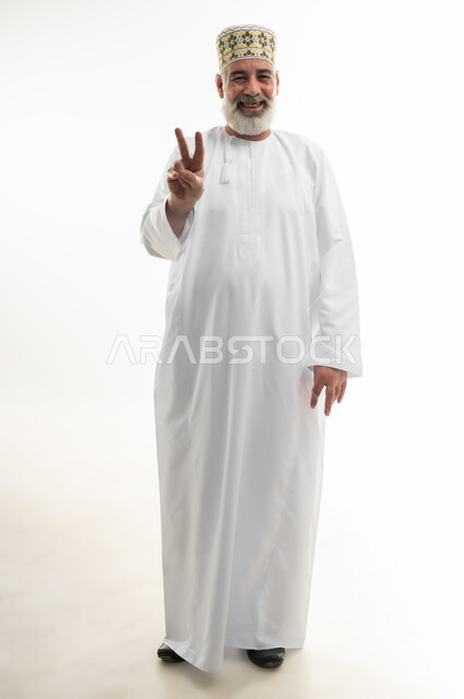 رفع أصابع اليد بعلامة النصر، الفوز والانتصار وتحقيق الأهداف، بورتريه لرجل كبير بالسن عربي خليجي عماني يرتدي الدشداشة والكمه ينظر إلى الكاميرا بإيماءات السعادة والسرور، الشعور بالقوة والرضا والسلام، الجسم بالطول الكامل، خلفية بيضاء