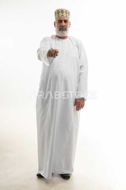 توجيه أصابع الاتهام إلى شخص ما، بورتريه لرجل كبير بالسن عربي خليجي عماني يرتدي الدشداشة والكمه يشير باصبع السبابة نحو الكاميرا بشكل مباشر بإيماءات الجدية والغضب، الإشارة إلى مكان ما محدد، الجسم بالطول الكامل، خلفية بيضاء