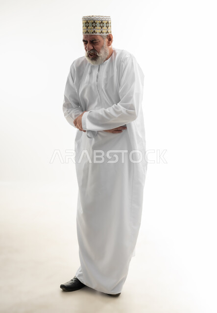 أهمية زيارة الطبيب والحصول على قسط من الراحة، إمساك البطن والشعور بآلام المغص، بورتريه لرجل كبير بالسن عربي خليجي عماني يرتدي الدشداشة والكمه يضع يديه على معدته بإيماءات الاضطراب والانزعاج، ضرورة أخذ الدواء للتعافي من المرض،  الجسم بالطول الكامل، خلفية بيضاء
