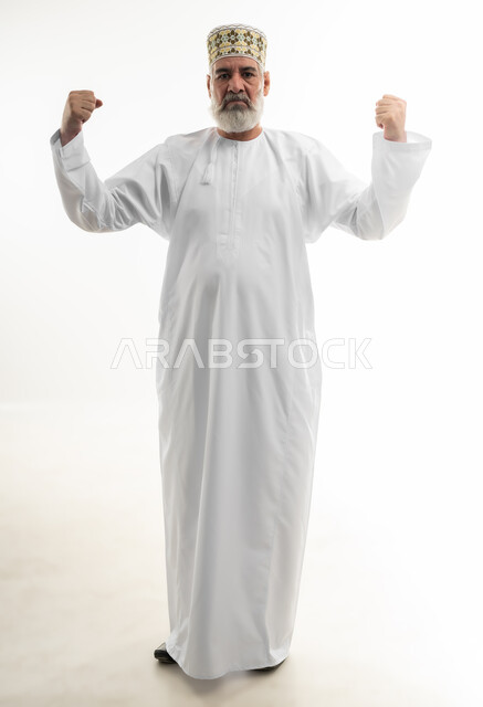 رفع اليدين لإظهار القوة وعرض العضلات، بورتريه لرجل كبير بالسن عربي خليجي عماني يرتدي الدشداشة والكمه ينظر إلى الكاميرا بإيماءات الجدية، ممارسة الرياضة بشكل منتظم للحفاظ على جسم صحي، التمتع باللياقة وبنية جسدية قوية، الجسم بالطول الكامل، خلفية بيضاء