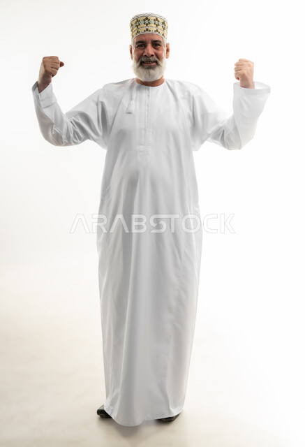 رفع اليدين لإظهار القوة وعرض العضلات، بورتريه لرجل كبير بالسن عربي خليجي عماني يرتدي الدشداشة والكمه ينظر إلى الكاميرا بإيماءات الجدية، ممارسة الرياضة بشكل منتظم للحفاظ على جسم صحي، التمتع باللياقة وبنية جسدية قوية، الجسم بالطول الكامل، خلفية بيضاء