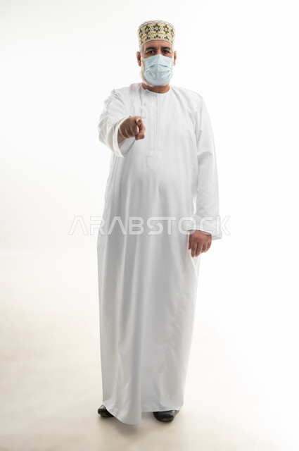 اتباع الإجراءات الاحترازية لمنع انتشار العدوى، بورتريه لرجل كبير بالسن عربي خليجي عماني يرتدي الدشداشة والكمه يضع الكمامة الطبية على وجهه ويشير باصبع السبابة نحو الكاميرا، اتخاذ التدابير اللازمة للوقاية من فيروس كورونا، الجسم بالطول الكامل، خلفية بيضاء