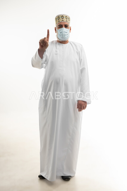 اتباع الإجراءات الاحترازية لمنع انتشار العدوى، بورتريه لرجل كبير بالسن عربي خليجي عماني يرتدي الدشداشة والكمه يضع الكمامة الطبية على وجهه ويرفع اصبع السبابة نحو الأعلى، اتخاذ التدابير اللازمة للوقاية من فيروس كورونا، الجسم بالطول الكامل، خلفية بيضاء